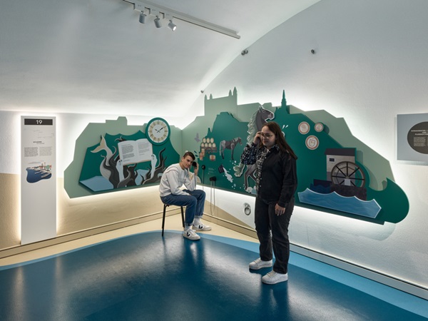 Zwei Jugendliche an Hör-Station im Donauschwäbischen Museum in Ulm. Im Hintergrund vereinfachte Darstellung einer historischen Stadt am Fluss